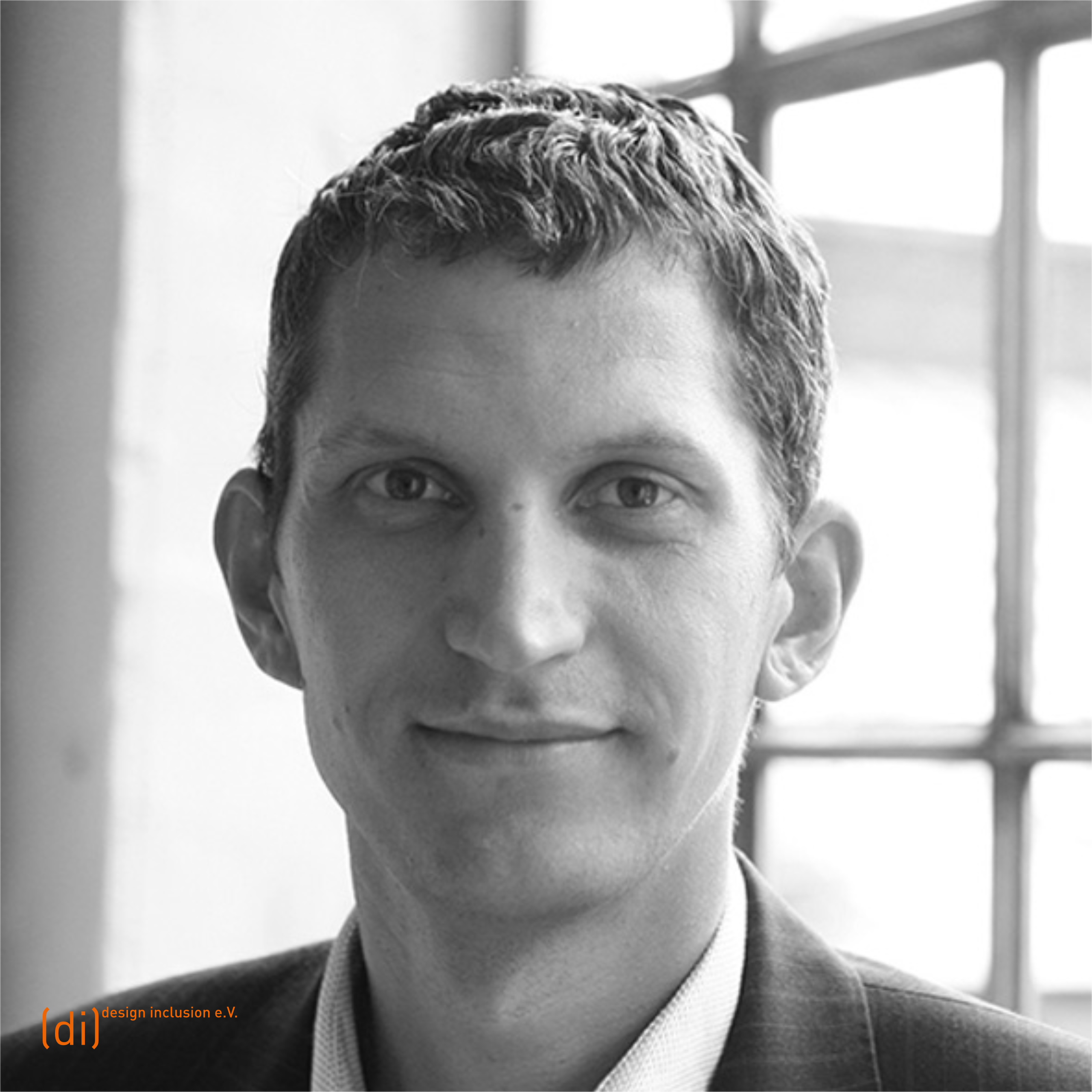 Frontales Porträt von Mathias Knigge mit kurzem, gewelltem Haar. Er trägt ein dunkles Jackett mit feinem Streifen und ein helles Hemd und schaut freundlich in die Kamera. Im Hintergrund ist ein großen Fenster mit mehreren rechteckigen Scheiben.