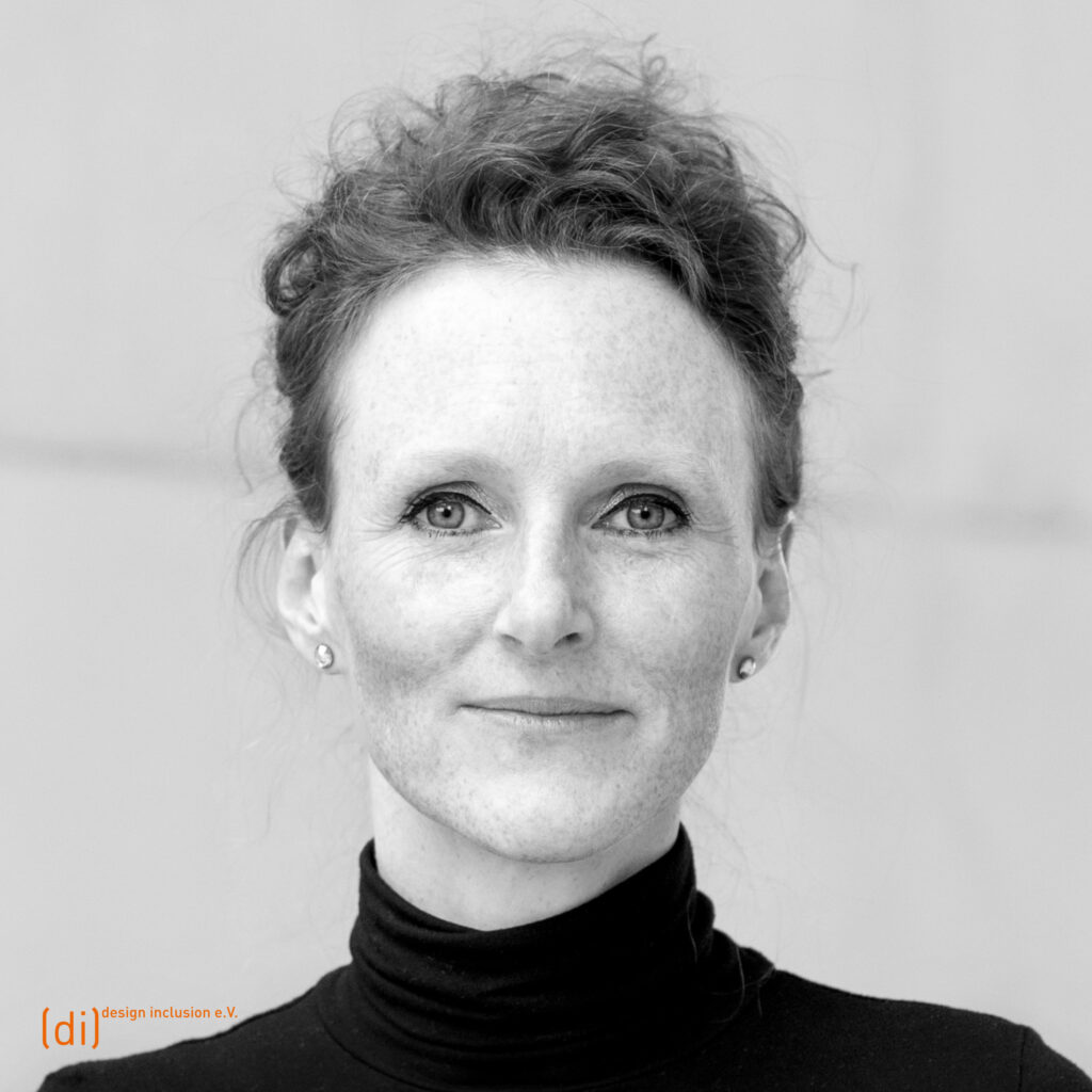 Frontales Porträt von Professorin Carlin Schreiber mit lockigem, hochgestecktem Haar. Sie trägt einen schwarzen Rollkragenpullover und schaut direkt in die Kamera. Der Hintergrund ist unscharf und fast einfarbig.