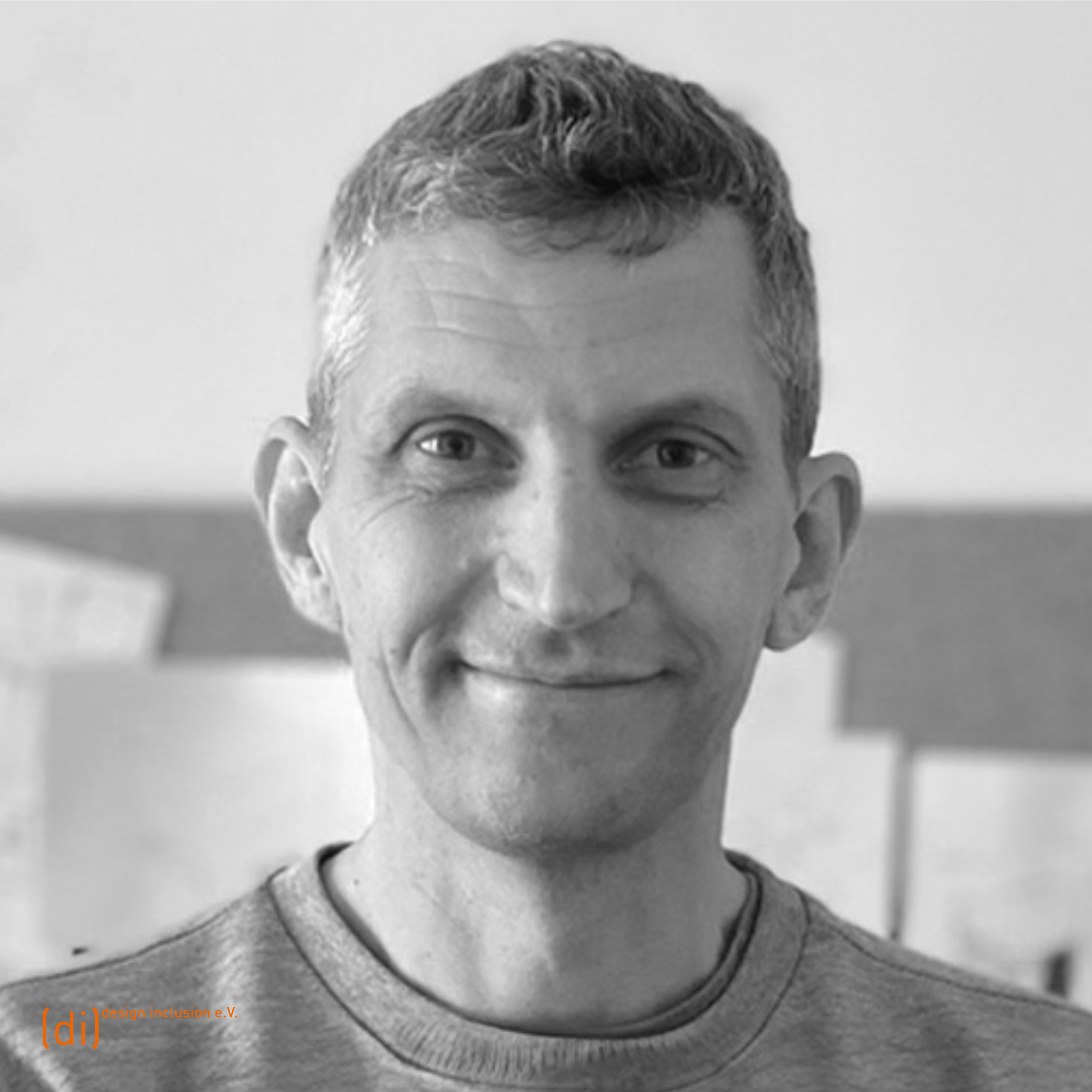 Porträt von Mathias Knigge mit kurzem Haar und einem grauen Pullover. Er steht vor einer Wand mit mehreren Papieren angespannt. Er lächelt freundlich in die Kamera.