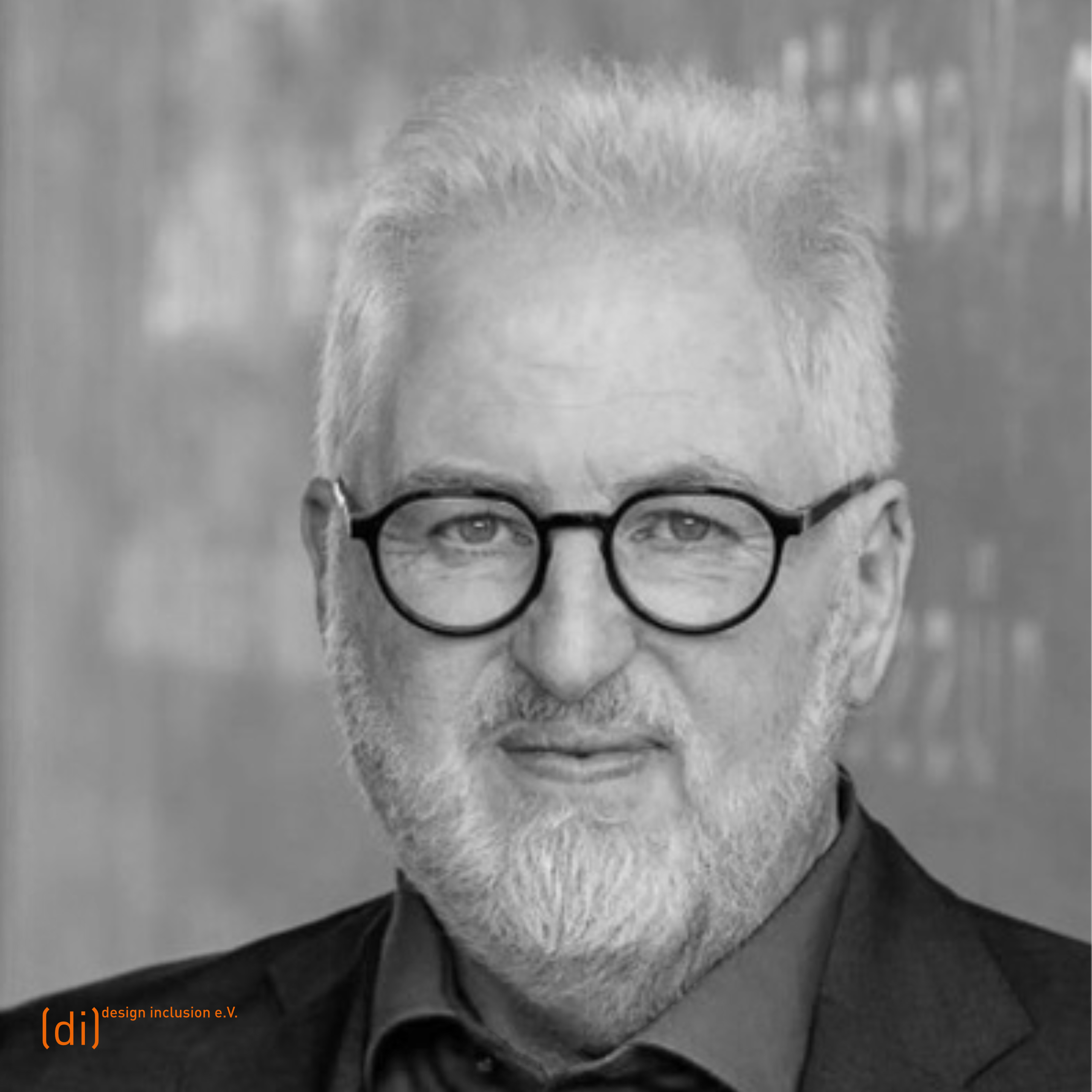 Porträt von Wolfgang Sattler mit kurzem, weißem Haar und Bart. Er trägt eine dunkle Brille und ein dunkles Jackett über einem Hemd. Sein Blick ist freundlich in die Kamera gerichtet. Im Hintergrund ist eine unscharfe Wand mit grafischen Elementen und Schriftzügen zu erkennen.